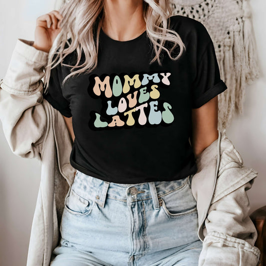 Graphic tee for women. The color of the shirt is black. In a colorful, typically 70s retro-style font it says Mommy Loves Lattes.