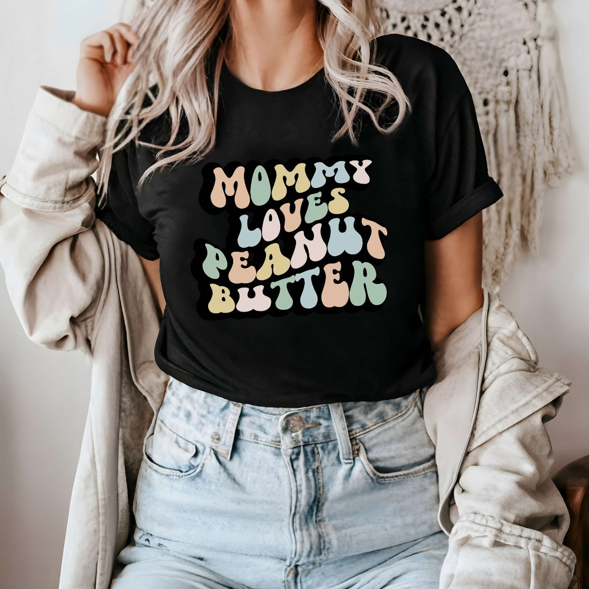 Graphic tee for women. The color of the shirt is black. In a colorful, typically 70s retro-style font it says Mommy Loves Peanut Butter.