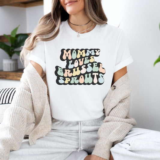 Graphic tee for women. The color of the shirt is white. In a colorful, typically 70s retro-style font it says Mommy Loves Brussel Sprouts.