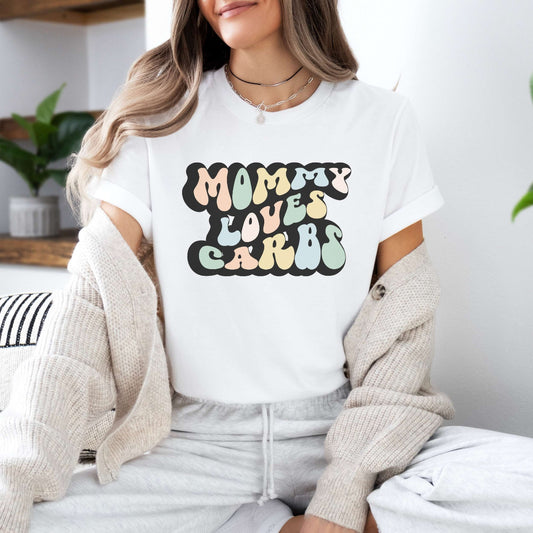 Graphic tee for women. The color of the shirt is white. In a colorful, typically 70s retro-style font it says Mommy Loves Carbs.