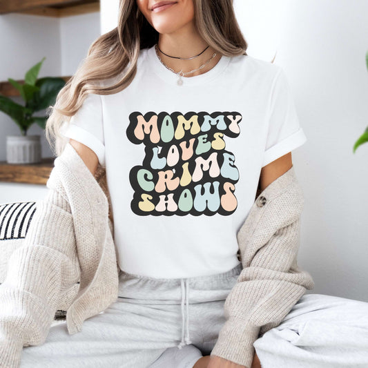 Graphic tee for women. The color of the shirt is white. In a colorful, typically 70s retro-style font it says Mommy Loves Crime Shows.