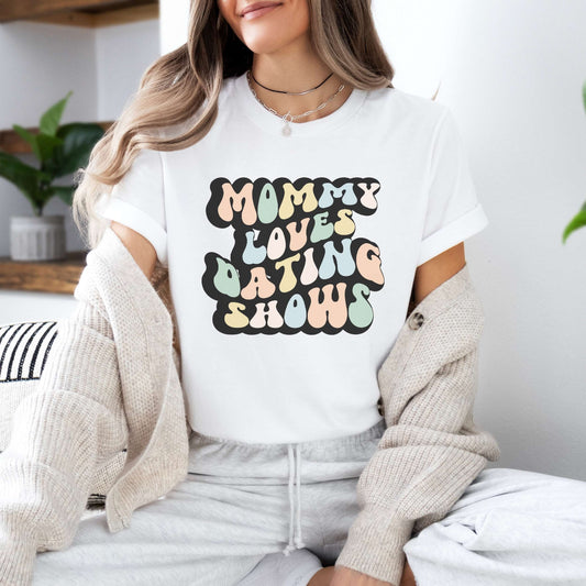 Graphic tee for women. The color of the shirt is white. In a colorful, typically 70s retro-style font it says Mommy Loves Dating Shows.