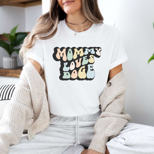 Graphic tee for women. The color of the shirt is white. In a colorful, typically 70s retro-style font it says Mommy Loves DOGE.