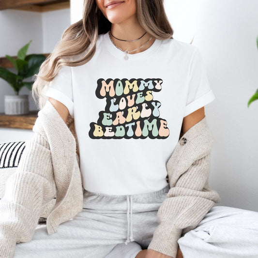 Graphic tee for women. The color of the shirt is white. In a colorful, typically 70s retro-style font it says Mommy Loves Early Bedtime.