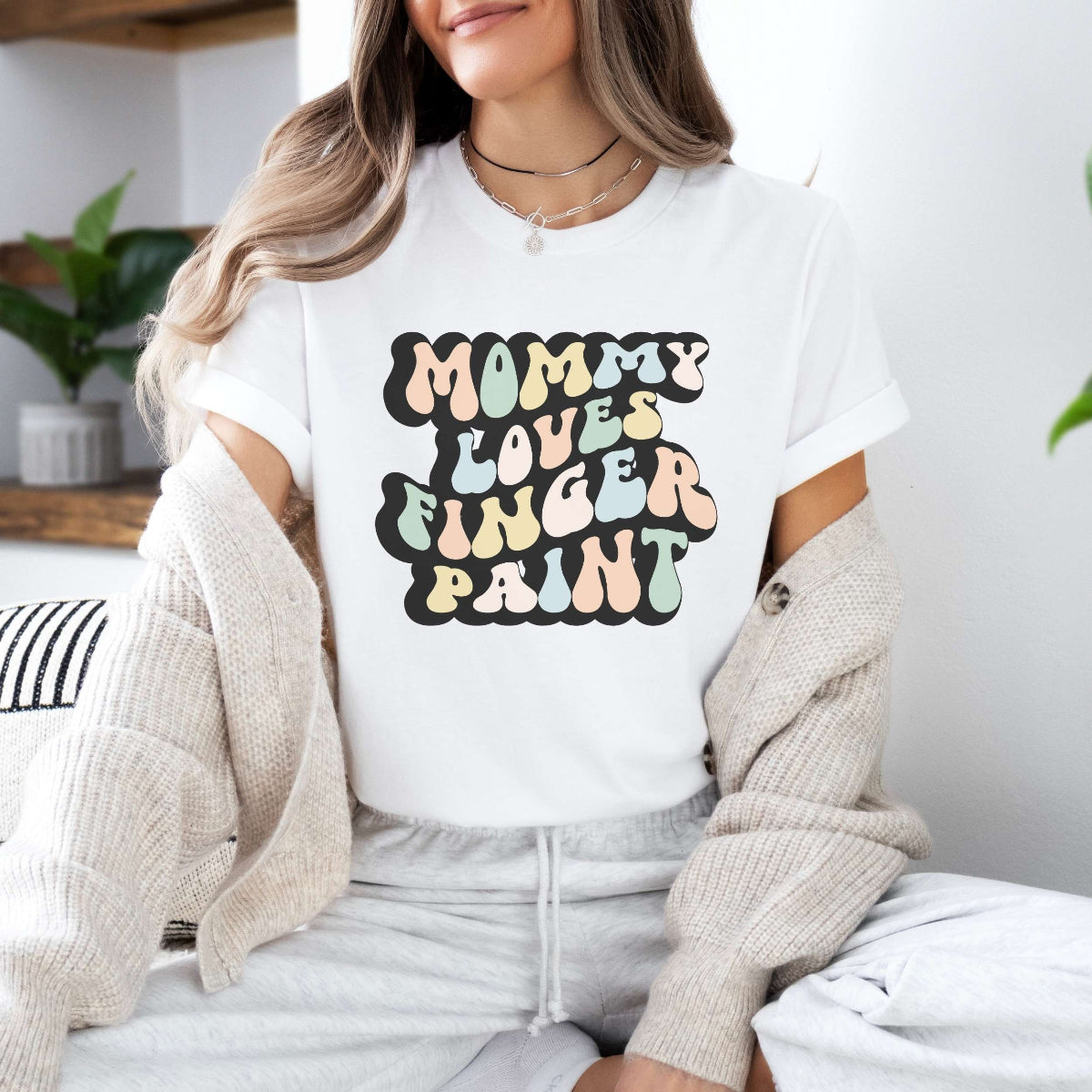 Graphic tee for women. The color of the shirt is white. In a colorful, typically 70s retro-style font it says Mommy Loves Finger Paint.
