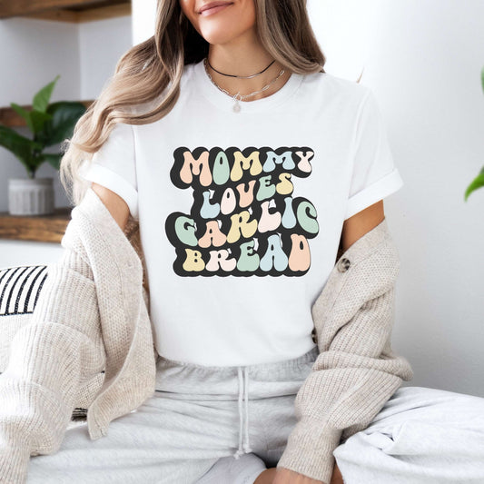 Graphic tee for women. The color of the shirt is white. In a colorful, typically 70s retro-style font it says Mommy Loves Garlic Bread.
