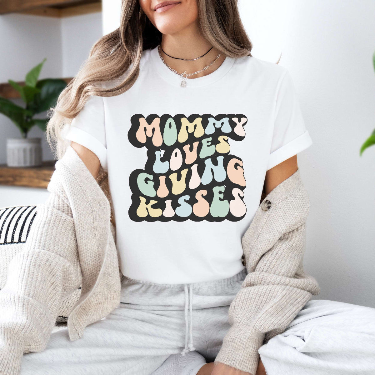 Graphic tee for women. The color of the shirt is white. In a colorful, typically 70s retro-style font it says Mommy Loves Giving Kisses.