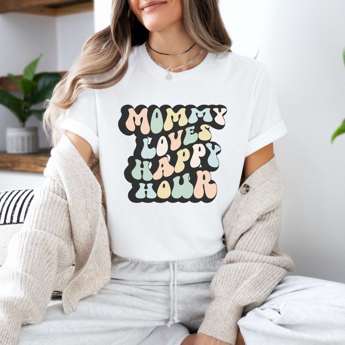 Graphic tee for women. The color of the shirt is white. In a colorful, typically 70s retro-style font it says Mommy Loves Happy Hour.