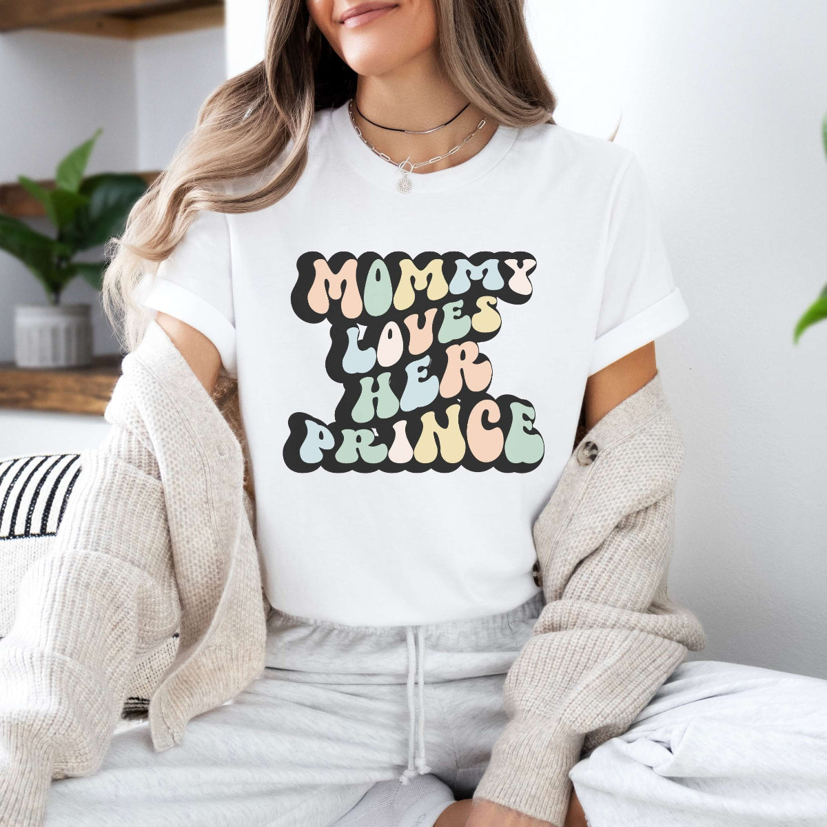 Graphic tee for women. The color of the shirt is white. In a colorful, typically 70s retro-style font it says Mommy Loves Her Prince.
