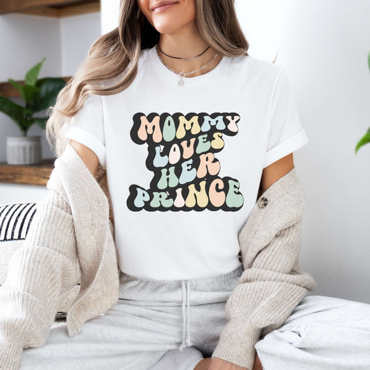 Graphic tee for women. The color of the shirt is white. In a colorful, typically 70s retro-style font it says Mommy Loves Her Prince.