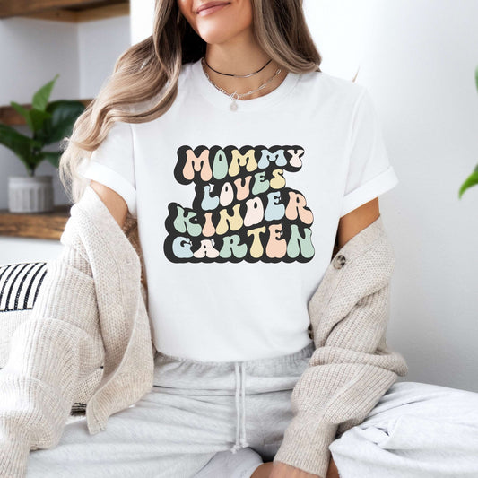 Graphic tee for women. The color of the shirt is white. In a colorful, typically 70s retro-style font it says Mommy Loves Kindergarten.