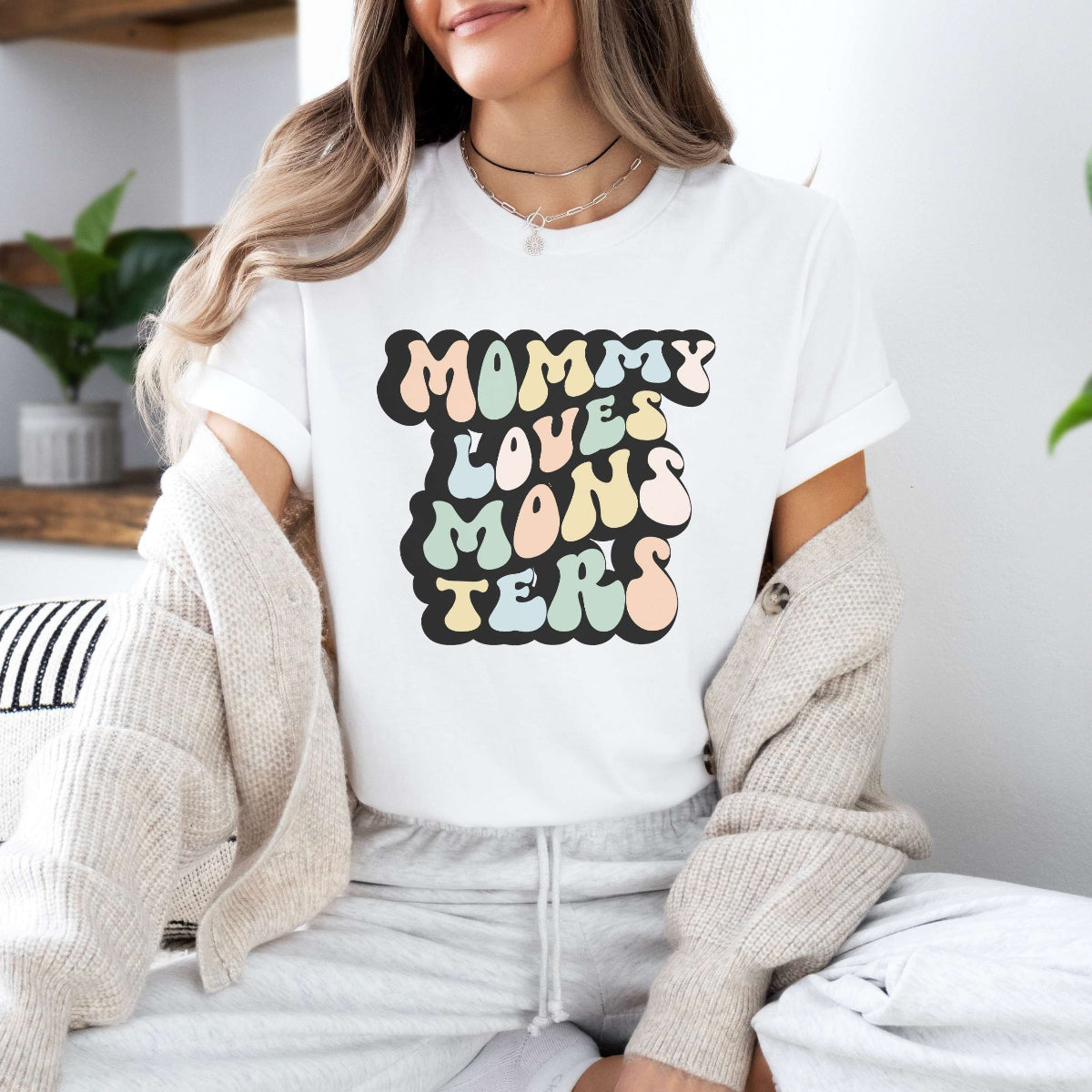 Graphic tee for women. The color of the shirt is white. In a colorful, typically 70s retro-style font it says Mommy Loves Monsters.
