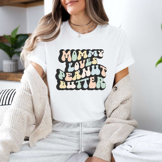 Graphic tee for women. The color of the shirt is white. In a colorful, typically 70s retro-style font it says Mommy Loves Peanut Butter.