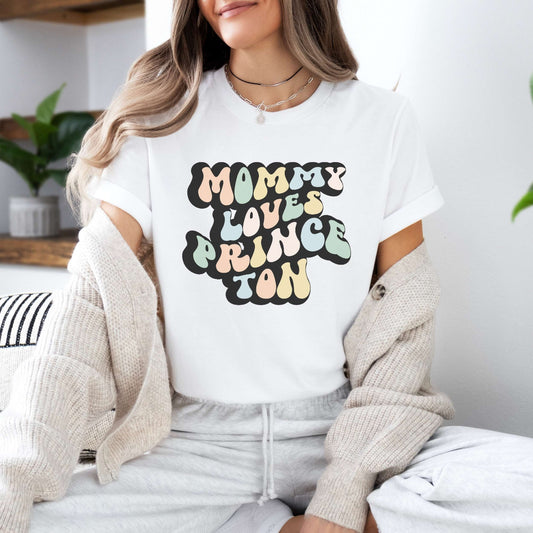 Graphic tee for women. The color of the shirt is white. In a colorful, typically 70s retro-style font it says Mommy Loves Princeton.