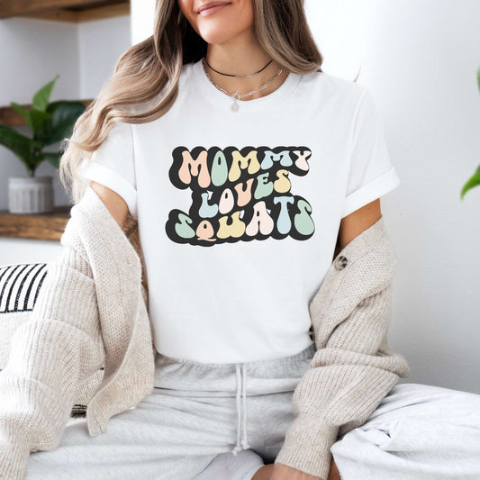 Graphic tee for women. The color of the shirt is white. In a colorful, typically 70s retro-style font it says Mommy Loves Squats.