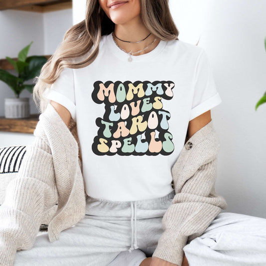 Graphic tee for women. The color of the shirt is white. In a colorful, typically 70s retro-style font it says Mommy Loves Tarot Spells.