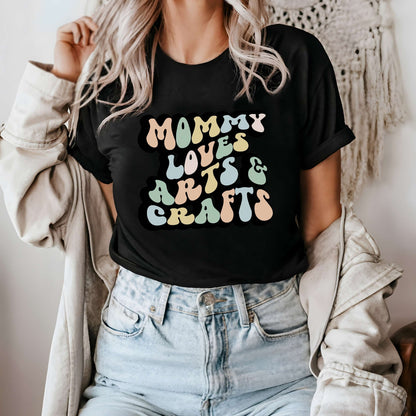 Graphic tee for women. The color of the shirt is black. In a colorful, typically 70s retro-style font it says Mommy Loves Arts & Crafts.