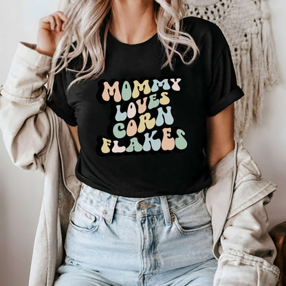 Graphic tee for women. The color of the shirt is black. In a colorful, typically 70s retro-style font it says Mommy Loves Corn Flakes.