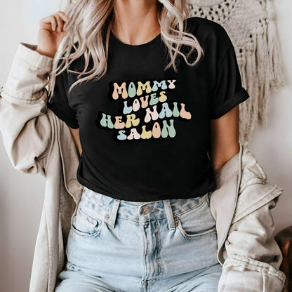 Graphic tee for women. The color of the shirt is black. In a colorful, typically 70s retro-style font it says Mommy Loves Her Nail Salon.