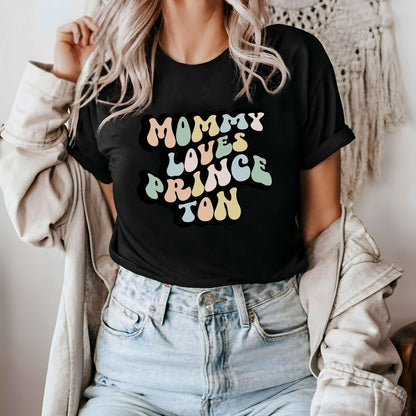 Graphic tee for women. The color of the shirt is black. In a colorful, typically 70s retro-style font it says Mommy Loves Princeton.