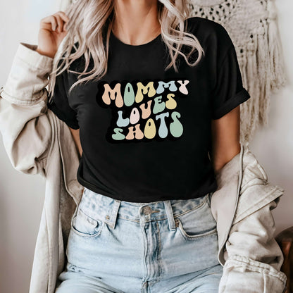 Graphic tee for women. The color of the shirt is black. In a colorful, typically 70s retro-style font it says Mommy Loves Shots.