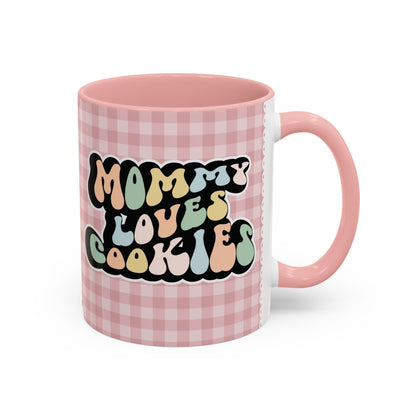 Perfect gift for mom. A decorative, 11oz ceramic mug. It’s wrapped in a soft pink-white checkered background. In a happy and colorful 70s retro-style font it says Mommy Loves Cookies, on both sides. 