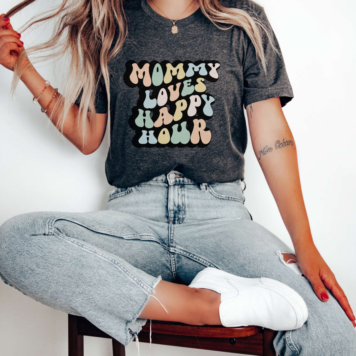 Graphic tee for women. The color of the shirt is gray heather. In a colorful, typically 70s retro-style font it says Mommy Loves Happy Hour.