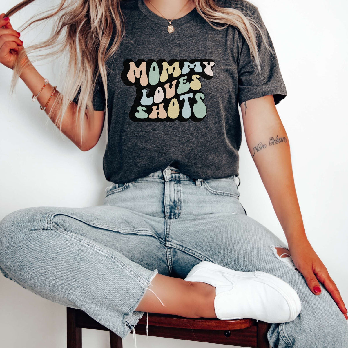 Graphic tee for women. The color of the shirt is gray heather. In a colorful, typically 70s retro-style font it says Mommy Loves Shots.