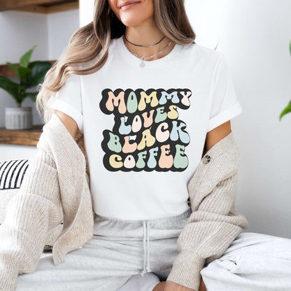 Graphic tee for women. The color of the shirt is white. In a colorful, typically 70s retro-style font it says Mommy Loves Black Coffee.