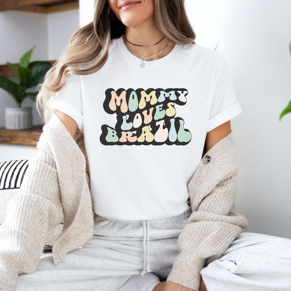 Graphic tee for women. The color of the shirt is white. In a colorful, typically 70s retro-style font it says Mommy Loves Brazil.