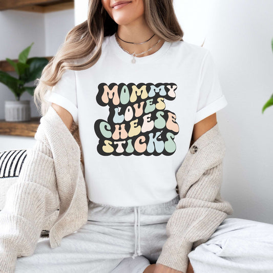 Graphic tee for women. The color of the shirt is white. In a colorful, typically 70s retro-style font it says Mommy Loves Cheese Sticks.