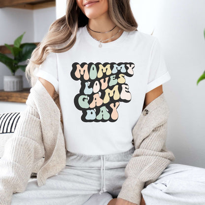 Graphic tee for women. The color of the shirt is white. In a colorful, typically 70s retro-style font it says Mommy Loves Game Day.