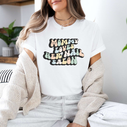 Graphic tee for women. The color of the shirt is white. In a colorful, typically 70s retro-style font it says Mommy Loves Her Nail Salon.