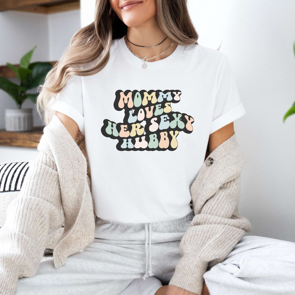Graphic tee for women. The color of the shirt is white. In a colorful, typically 70s retro-style font it says Mommy Loves her Sexy Hubby.