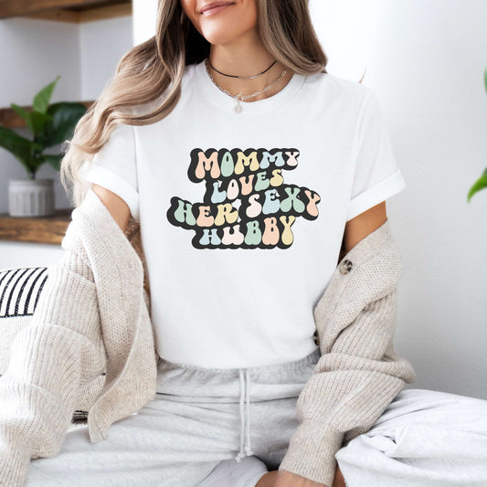 Graphic tee for women. The color of the shirt is white. In a colorful, typically 70s retro-style font it says Mommy Loves her Sexy Hubby.