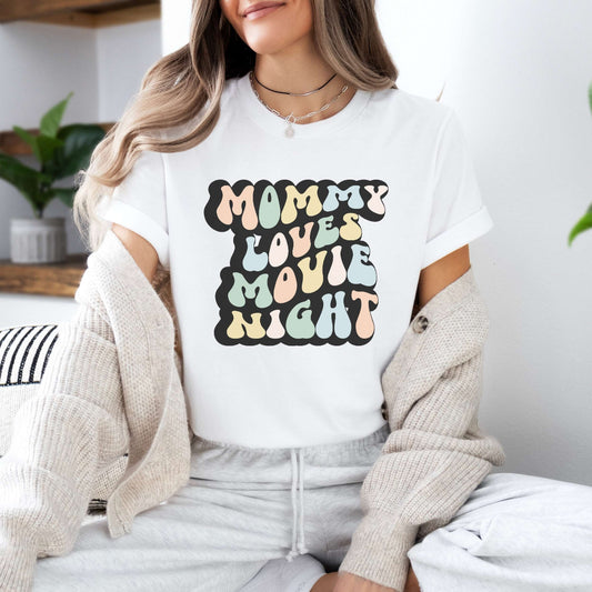 Graphic tee for women. The color of the shirt is white. In a colorful, typically 70s retro-style font it says Mommy Loves Movie Night.
