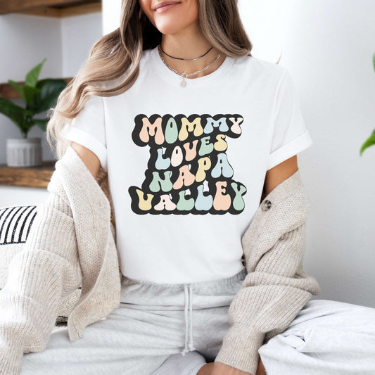 Graphic tee for women. The color of the shirt is white. In a colorful, typically 70s retro-style font it says Mommy Loves Napa Valley.