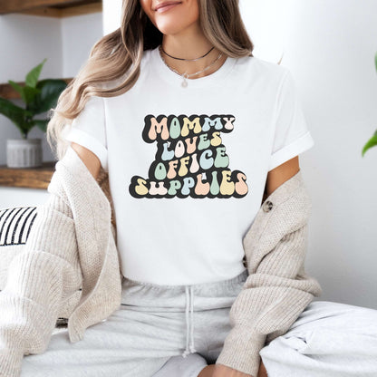 Graphic tee for women. The color of the shirt is white. In a colorful, typically 70s retro-style font it says Mommy Loves Office Supplies.
