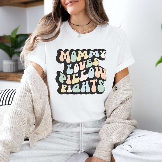 Graphic tee for women. The color of the shirt is white. In a colorful, typically 70s retro-style font it says Mommy Loves Pillow Fights.