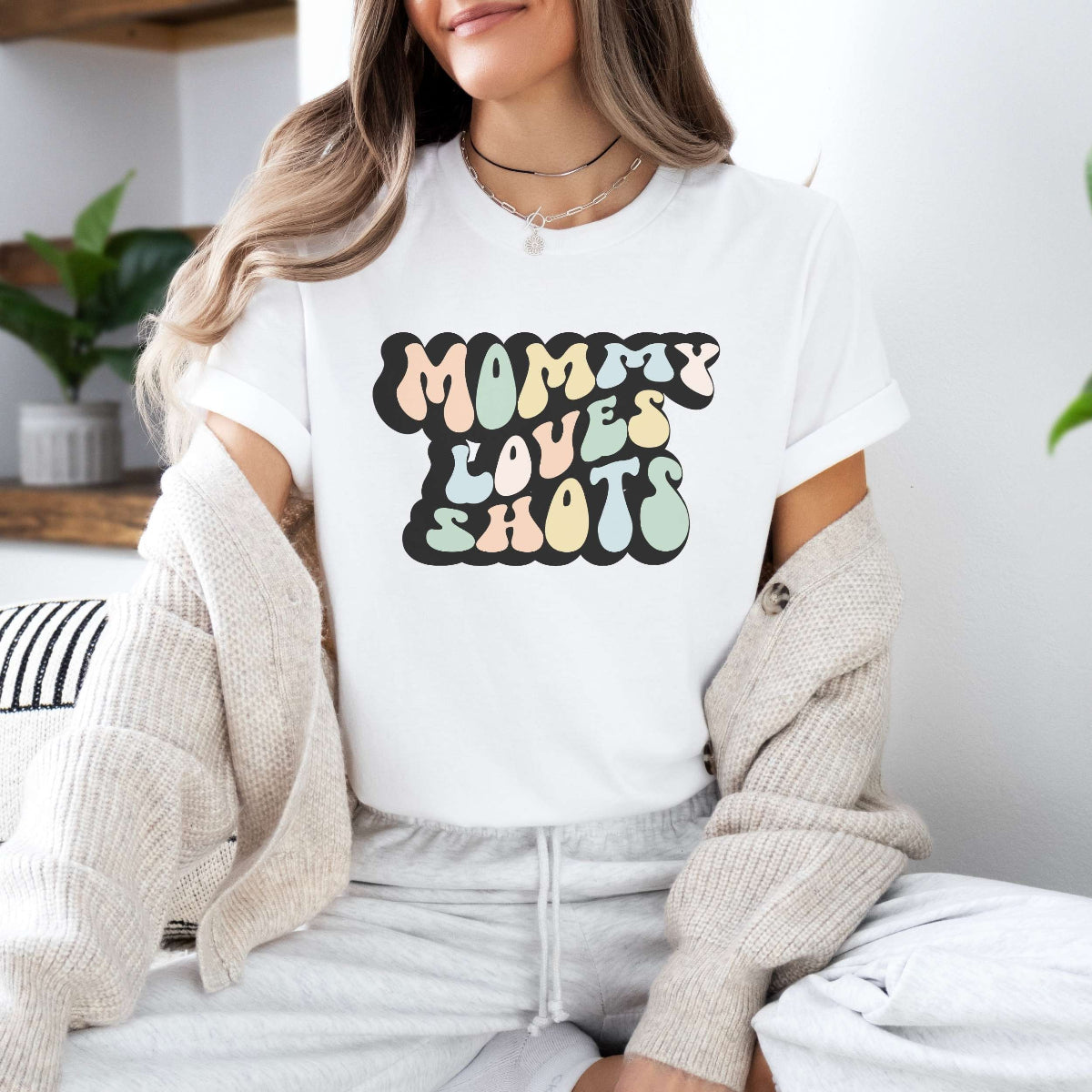 Graphic tee for women. The color of the shirt is white. In a colorful, typically 70s retro-style font it says Mommy Loves Shots.