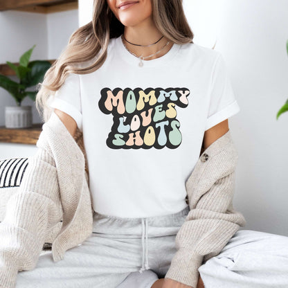Graphic tee for women. The color of the shirt is white. In a colorful, typically 70s retro-style font it says Mommy Loves Shots.