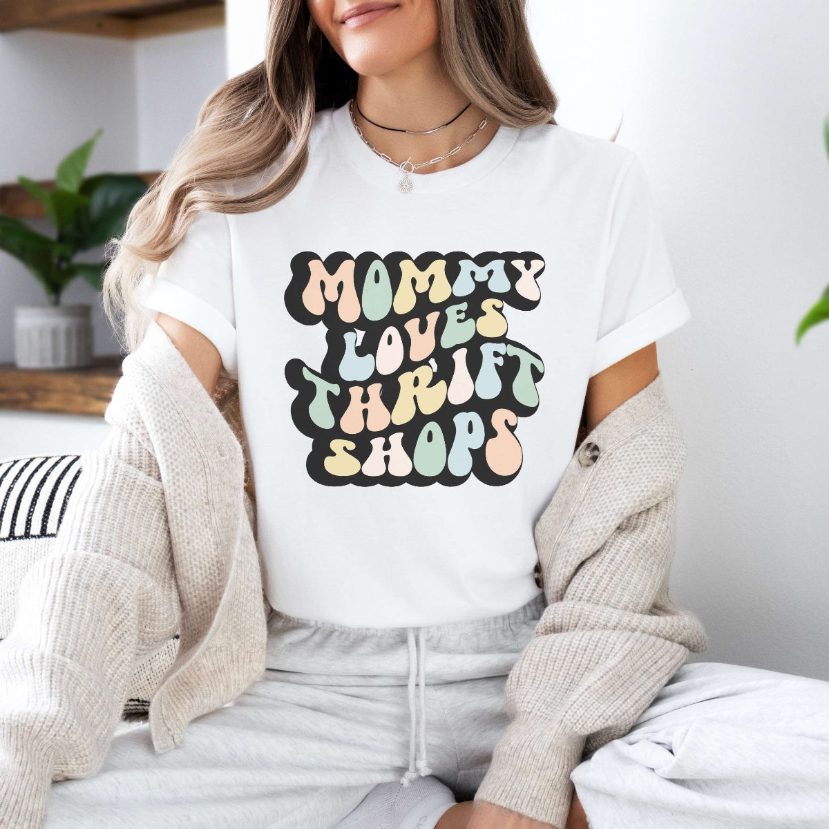 Graphic tee for women. The color of the shirt is white. In a colorful, typically 70s retro-style font it says Mommy Loves Thrift Shops.