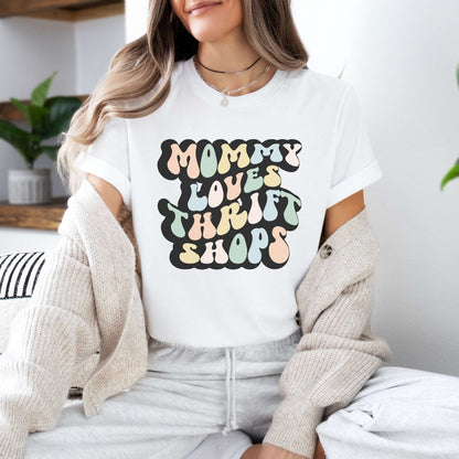 Graphic tee for women. The color of the shirt is white. In a colorful, typically 70s retro-style font it says Mommy Loves Thrift Shops.